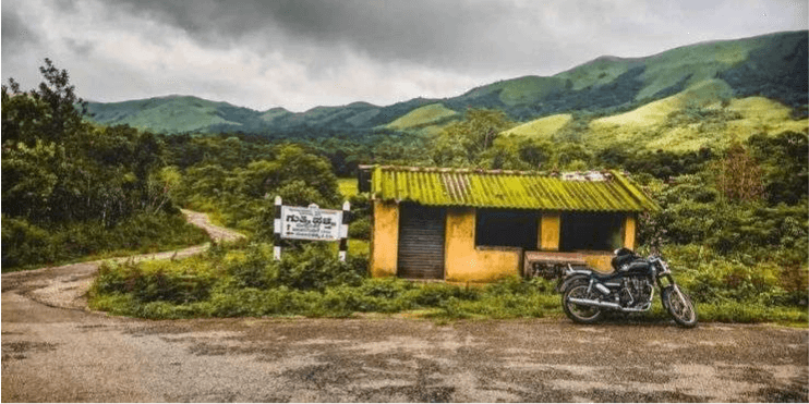 A motorcycle parked in front of a small shack in the tranquil village of Devaramane, surrounded by lush hills.