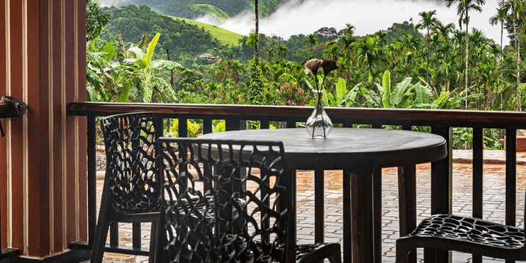A balcony with chairs and a table offers a scenic view of mountains, surrounded by lush hills and serene landscapes.