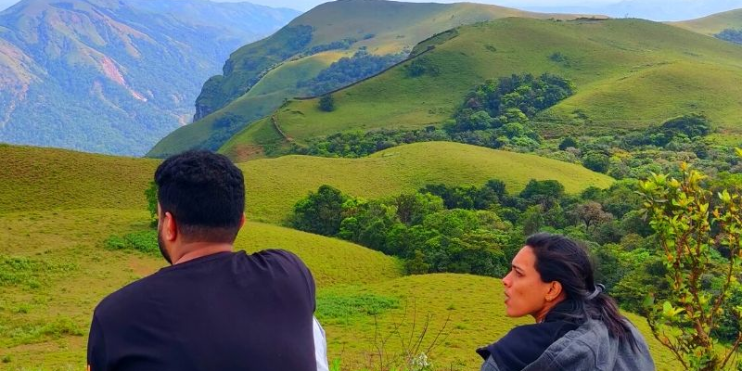 Two people sit on a hill, gazing at the valley below, with Ballarayanadurga Fort visible in the distance.