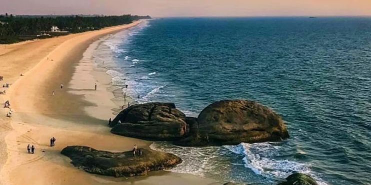 A serene beach in Karnataka with rocks and people strolling along the shore under light rain, capturing a peaceful monsoon vibe.