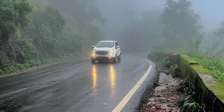 A white SUV drives along a foggy road, surrounded by lush greenery typical of Charmadi Ghat during the monsoon season.