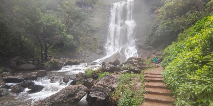 A man walks along a path beside a cascading waterfall, surrounded by lush greenery in Karnataka's scenic landscape.