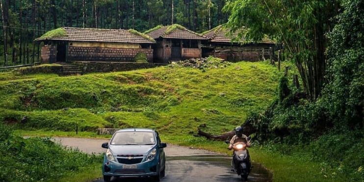 A motorcyclist rides down a road in front of a house, surrounded by greenery, on a sunny day.