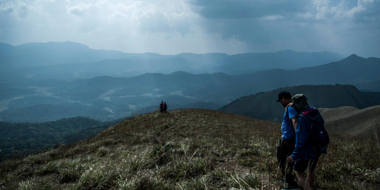Two hikers trek on a mountain trail under a cloudy sky, surrounded by lush greenery and the tranquility of nature.