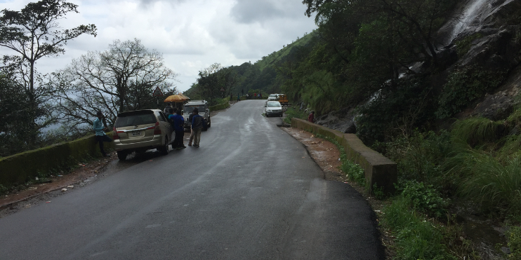 A scenic road in Charmadi Ghat with a car and two people walking, surrounded by lush greenery and mountainous terrain.