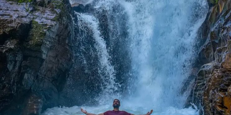 A man sits in the water at Manikyadhara Falls, surrounded by lush greenery and cascading water.