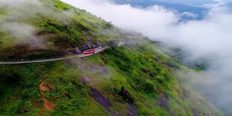 A red bus travels along a winding mountain road, surrounded by lush greenery and misty landscapes of Charmadi Ghat.