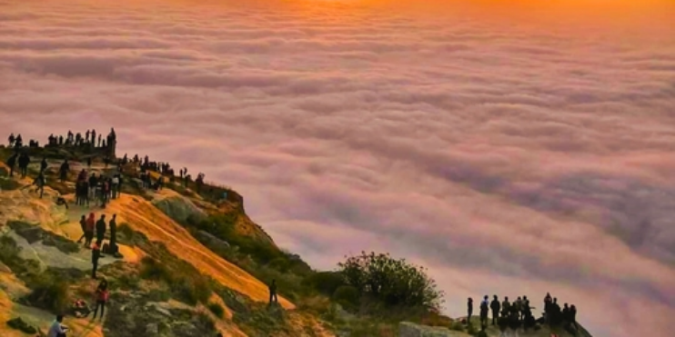 People standing on a mountain peak at sunset, overlooking a sea of clouds during the Skandagiri Trek near Bangalore.