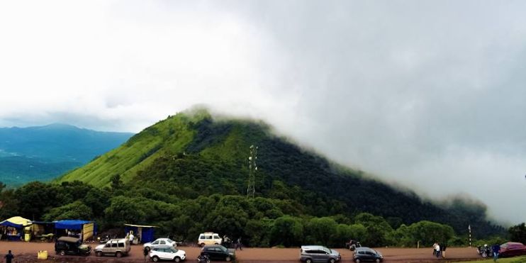 A mountain peak with several cars parked at the summit, surrounded by lush greenery typical of Karnataka's monsoon landscape.