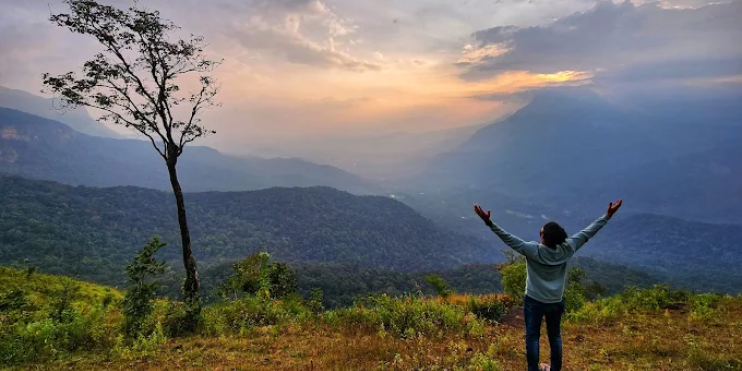 A man stands on a hill with arms outstretched, gazing at the mountains, embodying peace at Maidadi View Point.