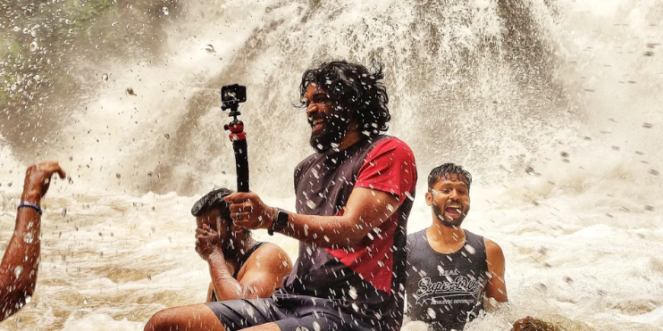 A group of people sitting in a shallow pool at Kodige Falls, taking pictures and enjoying the refreshing water.